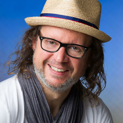 Auf der Fotografie ist Andreas Brichta zu sehen. Ein Mann mit lockigem Haar, einer Brille und einem Strohhut vor einer blauen Wand.