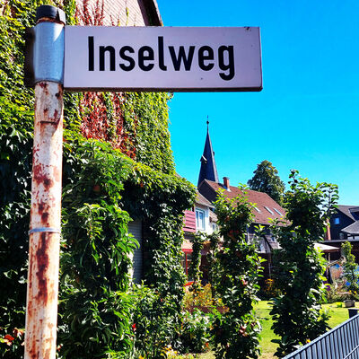 Ein Straßenschild mit dem Namen "Inselweg" vor dem Kirchturm in Sierße, Gemeinde Vechelde.